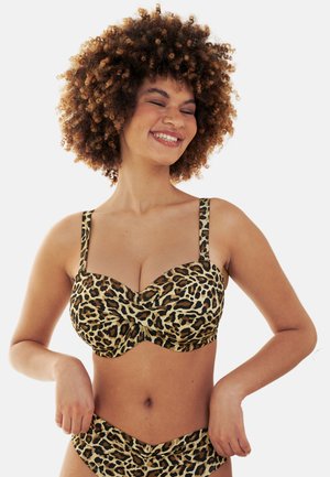Frau mit lockigem Haar, die einen Bikini mit Leopardenmuster trägt, lächelnd mit geschlossenen Augen und Händen auf der Hüfte vor hellem Hintergrund.
