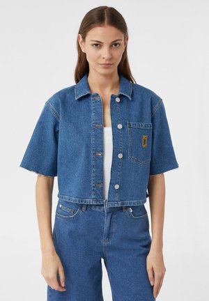 Cropped blue denim jacket with short sleeves, front buttons, and chest pocket featuring a yellow logo. Clean finish and no visible distressing.