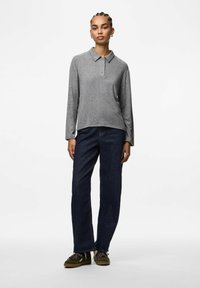 Mladá žena s pletencem vlasů, oblečená v šedém polokošili s dlouhým rukávem, tmavě modrých rovné džíny a zelených teniskách na šněrování, stojící na bílém pozadí.