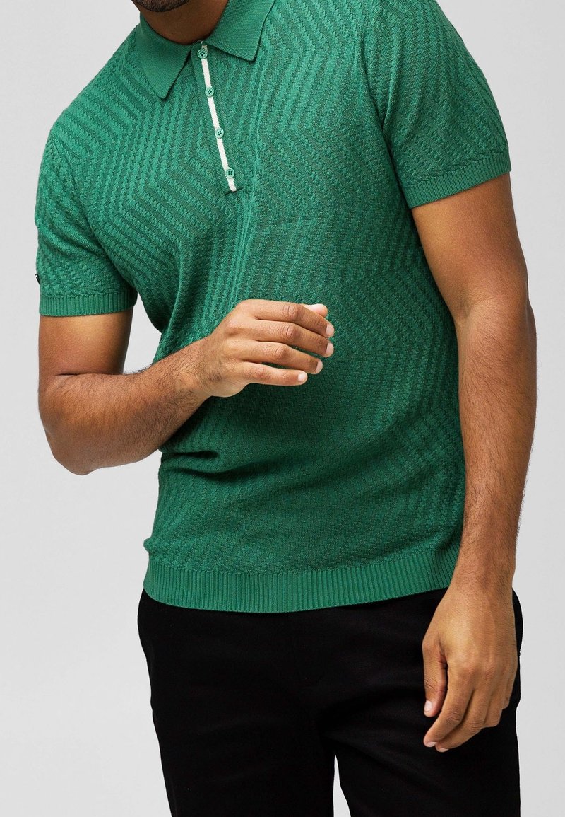 Homme portant un polo vert texturé à manches courtes avec boutons, assorti à un pantalon noir, sur un fond uni.
