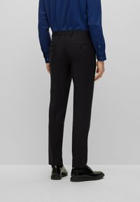 Pantaloni neri con una vestibilità sartoriale, tessuto liscio e due tasche posteriori. Indossati con una camicia da uomo blu a fantasia e scarpe nere.