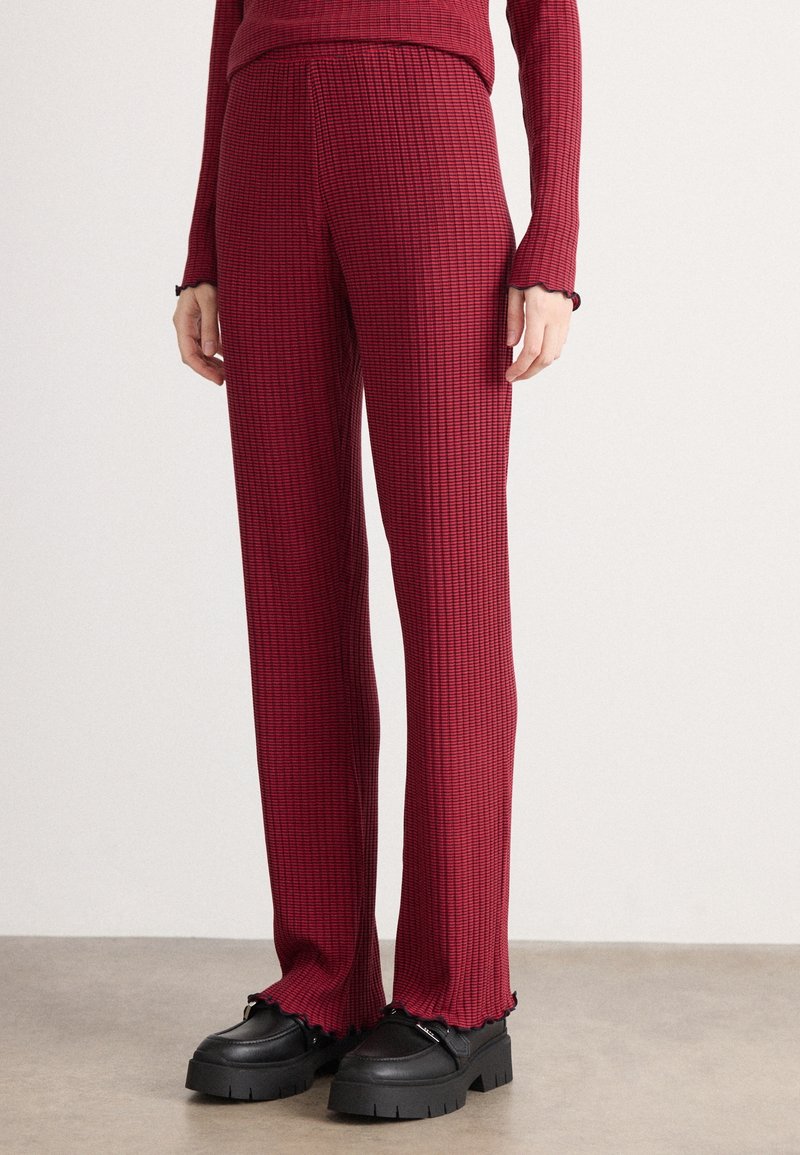 Femme portant un pantalon rouge texturé ajusté et un haut assorti avec des chaussures compensées noires épaisses sur un sol gris.