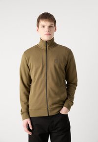 Oliven grønn zip-jakke laget av glatt stoff, høy krage, slim fit, med et firkantet logo på brystet og ribbede mansjetter. Sorte bukser brukt under.