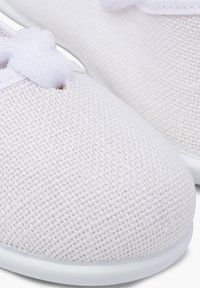 Close-up of white canvas shoes with a textured surface, featuring a small bow detail on the laces and a light rubber sole.