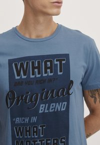 Blå bomulds t-shirt med korte ærmer og rund hals. Har store tekstgrafikker i forskellige skrifttyper, hovedsagelig i mørkeblå og lyseblå.