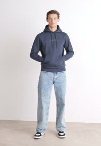 Marineblauwe hoodie met trekkoord, voorzak en lichtblauwe losse jeans. Schoeisel met zwart-witte accenten. Eenvoudig, casual ontwerp.
