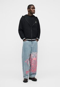 Άνδρας που στέκεται με τα χέρια στις τσέπες, φοράει μαύρο φούτερ με φερμουάρ, φαρδιά ανοιχτό μπλε τζιν με μεγάλο ροζ γραφικό στο δεξί πόδι και σκούρα παπούτσια.