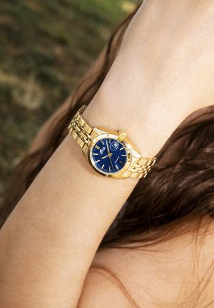 Montre-bracelet en or avec un cadran bleu, un bracelet métallique et un affichage de la date. Elle possède un boîtier rond et des surfaces texturées.