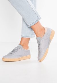 Graue Ledersneaker mit einem beigen Fersenstück und einer hellbeigen Gummisohle. Merkmale sind die runde Zehenform, das Low-Top-Design und die Schnürung.
