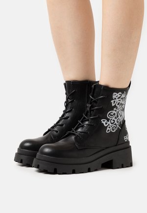 Bottes de cheville en cuir noir avec une semelle épaisse, un devant à lacets et un texte en style graffiti blanc sur le côté. Texture lisse, construction robuste.