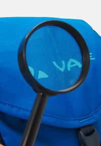 Mochila de tela azul con superficie texturizada. Una lupa negra enfoca el logo, revelando los finos detalles del material.