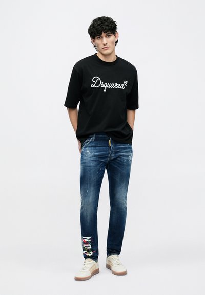 Μαύρο oversized t-shirt με το λογότυπο "Dsquared²", συνδυασμένο με μπλε τζιν με φθορές, πιτσιλιές χρώματος και πολύχρωμες λεπτομέρειες. Λευκά αθλητικά παπούτσια.