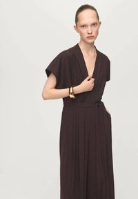 Ciemnobrązowa sukienka z krótkimi rękawami z naszywkami, głęboki dekolt w kształcie litery V, plisowany fason i pasek w talii. Złota bransoletka na nadgarstku.