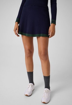 Modelo con una falda de punto azul marino con ribete verde, calcetines grises y zapatillas blancas, sobre un fondo de estudio liso.