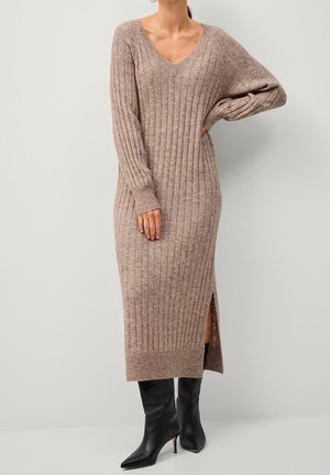 Femme portant une longue robe pull en maille côtelée taupe avec une fente latérale et des bottines à talons noires à bout pointu, debout, main sur la hanche.