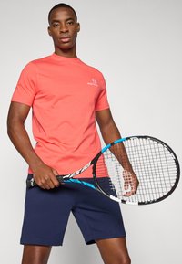 Koral kortermet skjorte laget av bomull, med en subtil logo. Marineblå shorts med løs passform. Personen holder et blått og svart tennisracket.