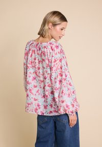 Blomstret bluse i let stof med pink og blå blomstermønstre. Har en sammensunken hals og voluminøse ærmer. Kombineret med blå jeans.