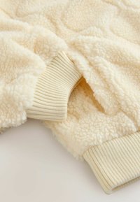 Tkanina boje kreme od sherpa s rebrastim manžetama. Mekana, teksturirana površina s ugodnim izgledom i suptilnim detaljima uzorka.
