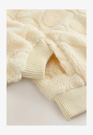 Tessuto sherpa color crema con polsini a costine. Superficie morbida e testurizzata con un aspetto accogliente e dettagli di motivo sottili.