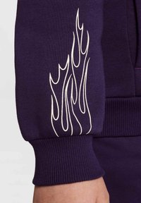 Sudadera morada con puños acanalados que presenta un diseño gráfico de llamas blancas en la manga izquierda. Textura suave y forma ajustada.