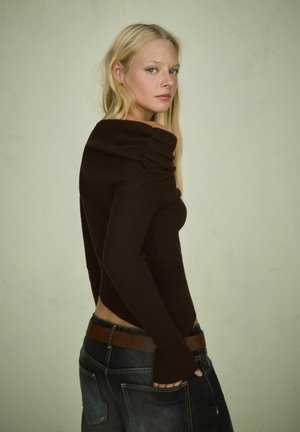 Donna bionda che indossa un maglione marrone a coste con spalle scoperte e jeans scuri con una cintura marrone, in piedi di lato contro uno sfondo neutro.