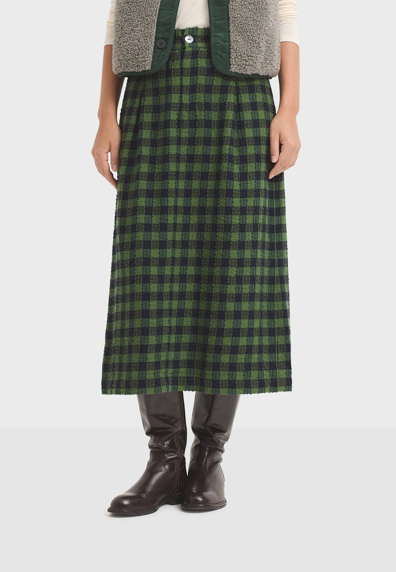 Falda midi de cuadros verdes y marinos, confeccionada en una tela texturizada, con pliegues y corte recto, combinada con botas altas de cuero marrón.