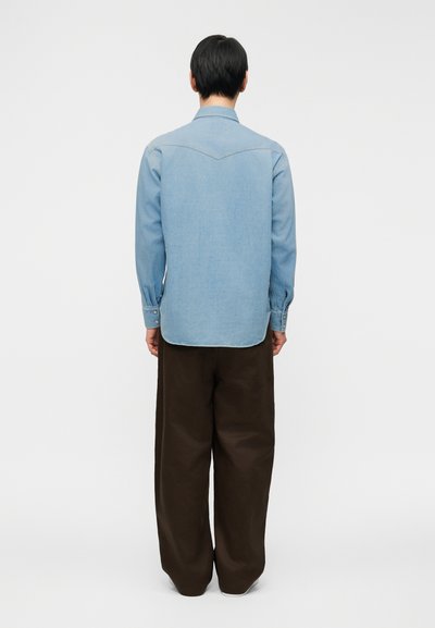 Ανοιχτό μπλε τζιν πουκάμισο με μακριά μανίκια και μυτερό γιακά, με καμπυλωτό τελείωμα και κουμπώματα, φορεμένο με σκούρο καφέ φαρδιά παντελόνια.