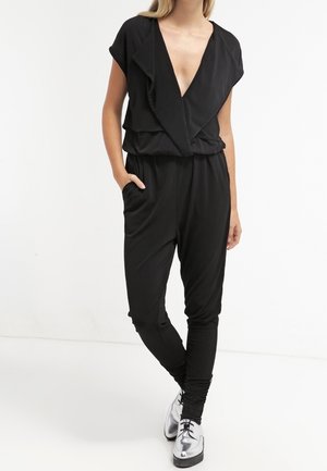 Kobieta w czarnym bezrękawnym kombinezonie z głębokim dekoltem w serek i falbanianym kołnierzem, stojąca boso na jednolitym białym tle.