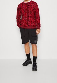Rood gemusterde trui met geribbelde manchetten, gecombineerd met zwarte shorts met twee vo pockets en logo-detail. Zwarte sneakers met veters.