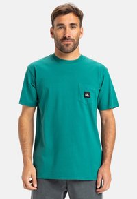 Tyrkysové bavlněné tričko s kulatým výstřihem, krátkými rukávy a kapsou na levé straně hrudníku s černobílou nášivkou.