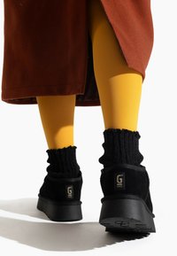 Botas de tobillo de ante negras con suelas de goma gruesas, combinadas con calcetines negros acanalados. Medias amarillas visibles y una prenda exterior marrón.