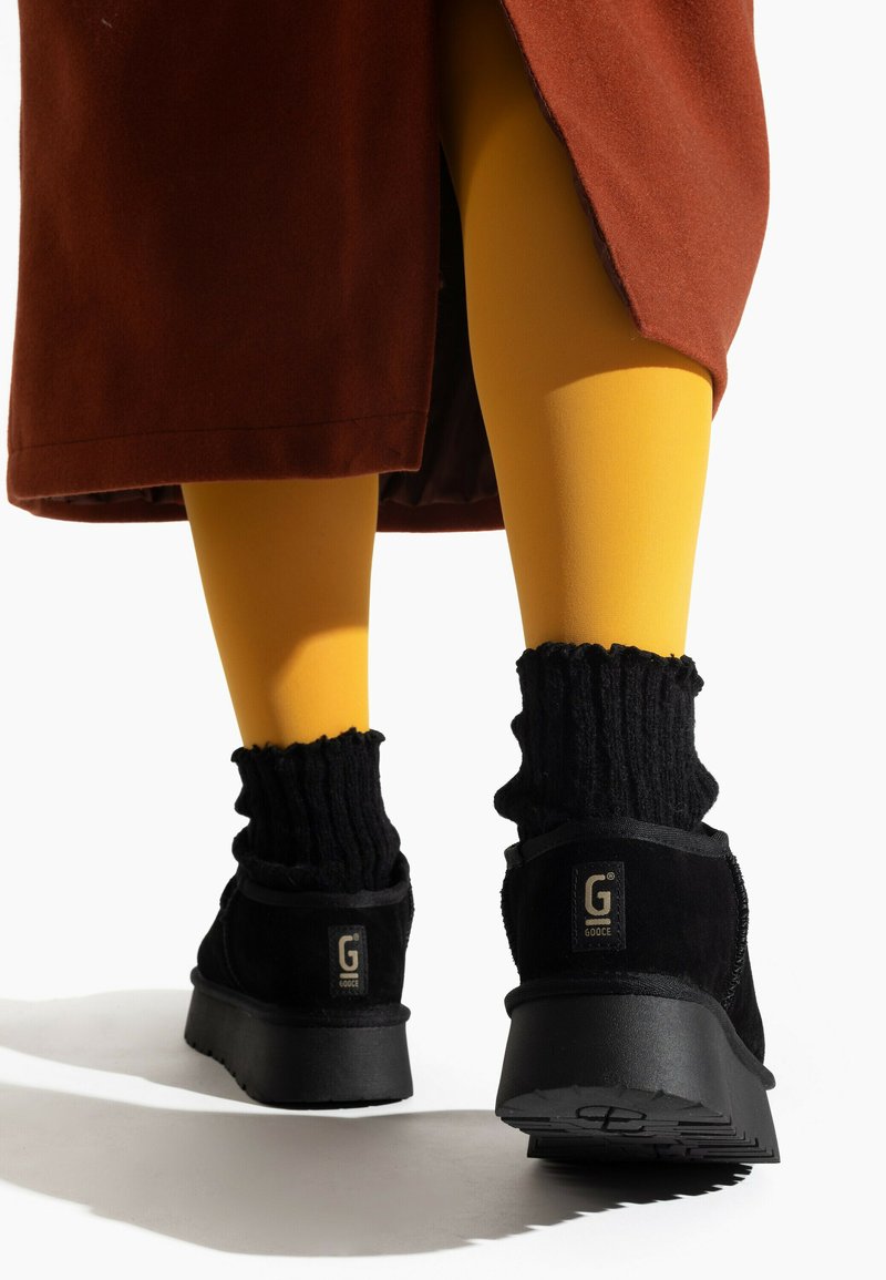 Botas de tobillo de ante negras con suelas de goma gruesas, combinadas con calcetines negros acanalados. Medias amarillas visibles y una prenda exterior marrón.