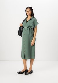 Abito verde con bottoni e cintura, maniche corte e due tasche frontali. Indossato con ballerine nere e una grande borsa nera.
