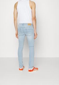 Vaqueros ajustados azul claro con un parche de cuero marrón con la etiqueta "ONLY & SONS" en la parte trasera, combinados con zapatillas deportivas naranjas y blancas y una camiseta sin mangas blanca.