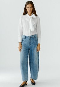 Witte blouse met een grote strik-kraag, gecombineerd met lichtblauwe denim jeans met wijde pijpen en zwarte puntige ballerina's. Minimalistisch ontwerp, zachte textuur.