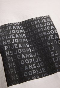 Witte stof met een zwarte patch waarop herhaaldelijk de tekst "JOOP! JEANS" in verschillende maten is afgebeeld. De patch heeft een gladde textuur.