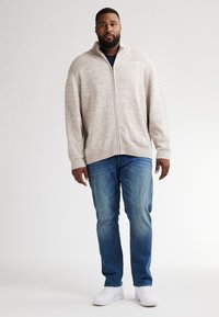 Světle šedý svetr na zip s ribbenými detaily, kombinovaný s modrými džíny a bílými teniskami. Tkanina vypadá měkce a strukturovaně.