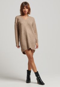 Vestido de punto beige con escote en V, corte relajado, hombros caídos y aberturas laterales, combinado con botas negras hasta el tobillo. Textura suave.