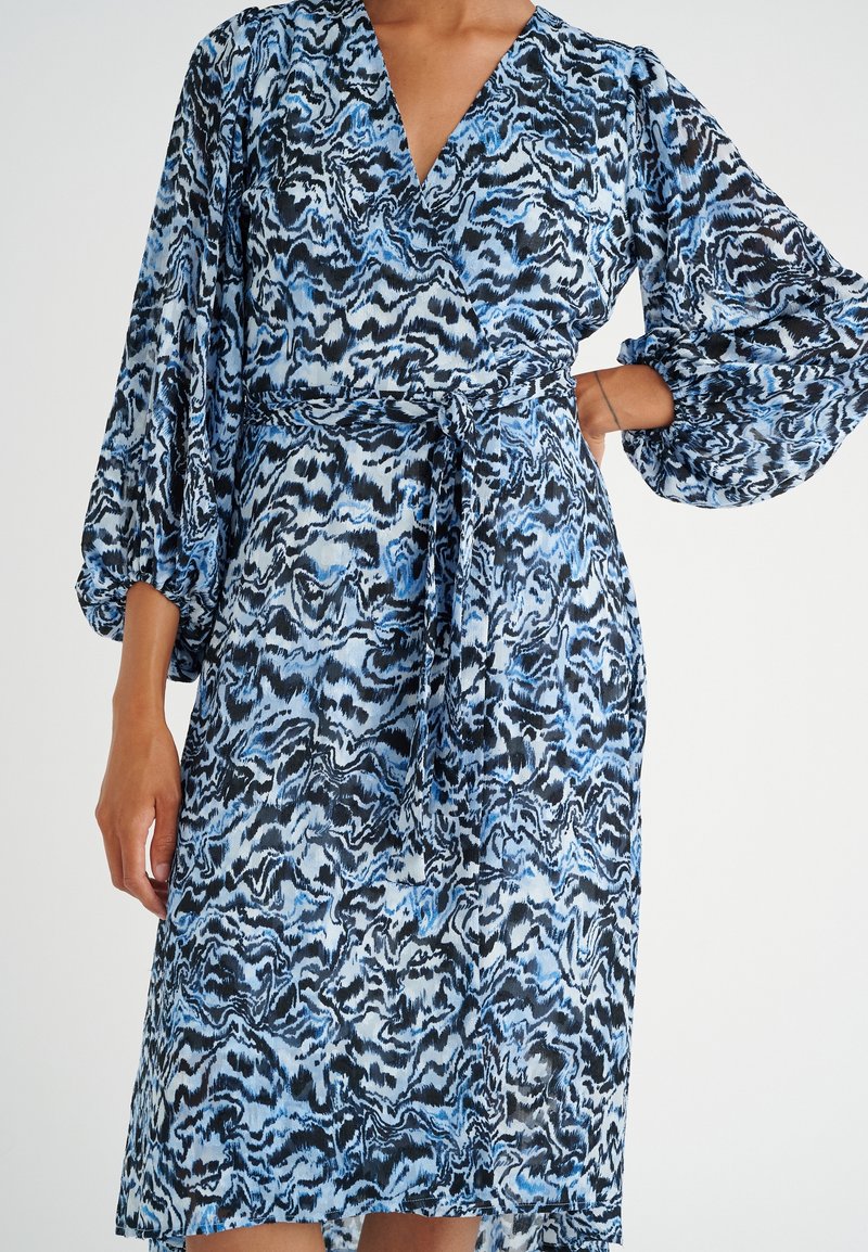 Blå och svart mönstrad klänning med långa puffärmar, ett knyt i midjan och en djup V-ringning. Lätt tyg med lätt lyster.