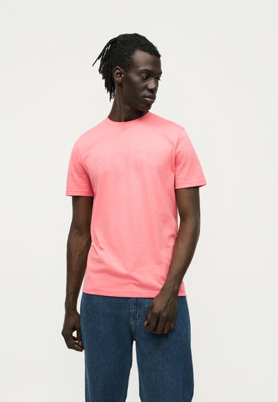 Homme avec des dreadlocks portant un T-shirt rose à manches courtes et un jean bleu, regardant vers le bas et vers sa gauche sur un fond uni.