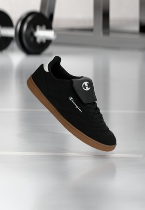 Schwarzer und brauner Champion-Sneaker schwebt in einem Fitnessstudio in der Luft, im Hintergrund ist eine verschwommene Langhantel zu sehen.