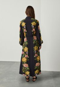 Abito lungo nero con ricami floreali vivaci in giallo, rosa e verde. Presenta un colletto e maniche lunghe, con una silhouette fluida.