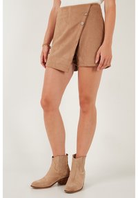 Shorts en daim beige avec un design croisé, dotés de deux boutons à l'avant et d'une texture lisse. Associés à des bottines beiges.