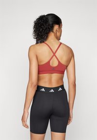 Bordeaux sportbeha met gekruiste banden; heeft een logo in het midden. Zwarte shorts met een brede tailleband waarop een herhaald logo-patroon staat.