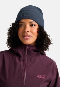 Donna con capelli ricci che indossa un cappello in pile blu navy di Jack Wolfskin e una giacca zip maroon di Jack Wolfskin, che guarda leggermente di lato.