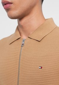 Barna cipzáras póló, texturált anyagból, bordás gallérral, és egy kis piros, fehér és kék logóval a mellkason.