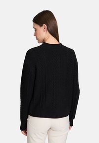 Mustan neulepuseron, jossa on herringbone-kuvio, varustettuna joustavaan kaulukseen ja pitkine hihoineen, päällä vaaleat housut.