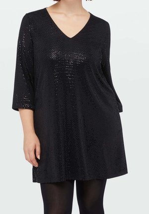 Frau trägt eine schwarze, langärmelige Pailletten-Tunika mit V-Ausschnitt, kombiniert mit schwarzen blickdichten Strumpfhosen, steht vor einem einfarbigen Hintergrund.