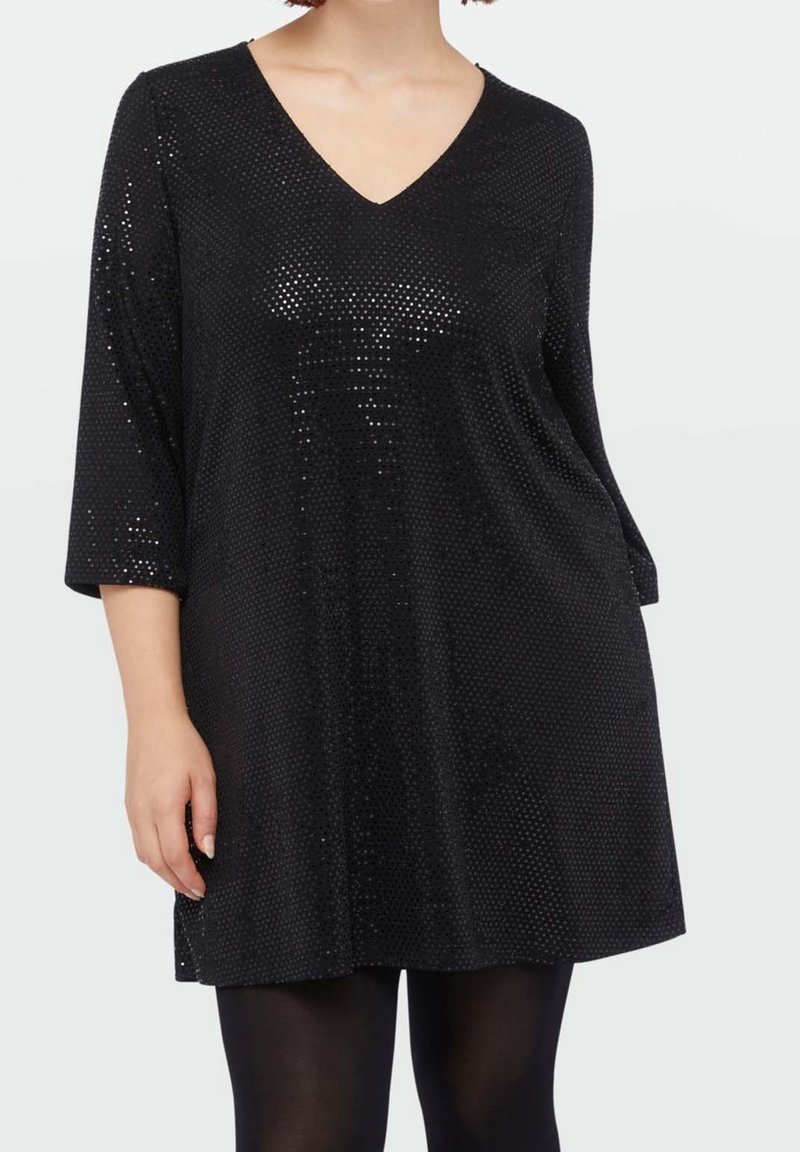 Femme portant une tunique noire à manches longues ornée de sequins avec un décolleté en V, associée à des collants opaques noirs, debout devant un fond uni.