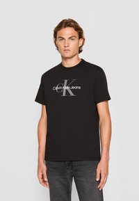 Camiseta de algodón negra con un gráfico central en blanco que dice "Calvin Klein Jeans" y un gran logo gris "CK". Cuello redondo clásico.
